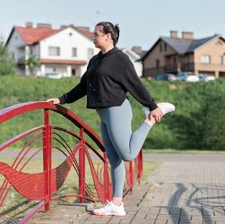 A woman in active wear performs a stretching exercise on a red bridge in a suburban park setting.